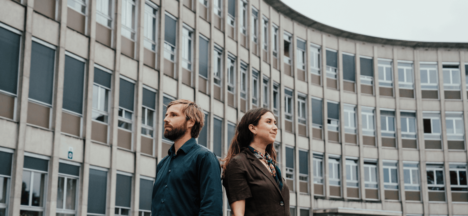 Thomas en Dayana staan rug aan rug voor de buitengevel van een campus.