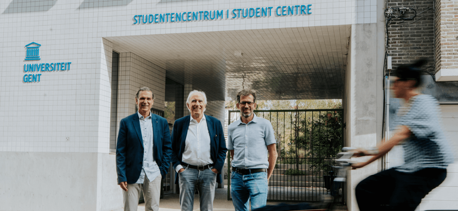 Drie formeel geklede mannen poseren voor het studentencentrum van UGent.