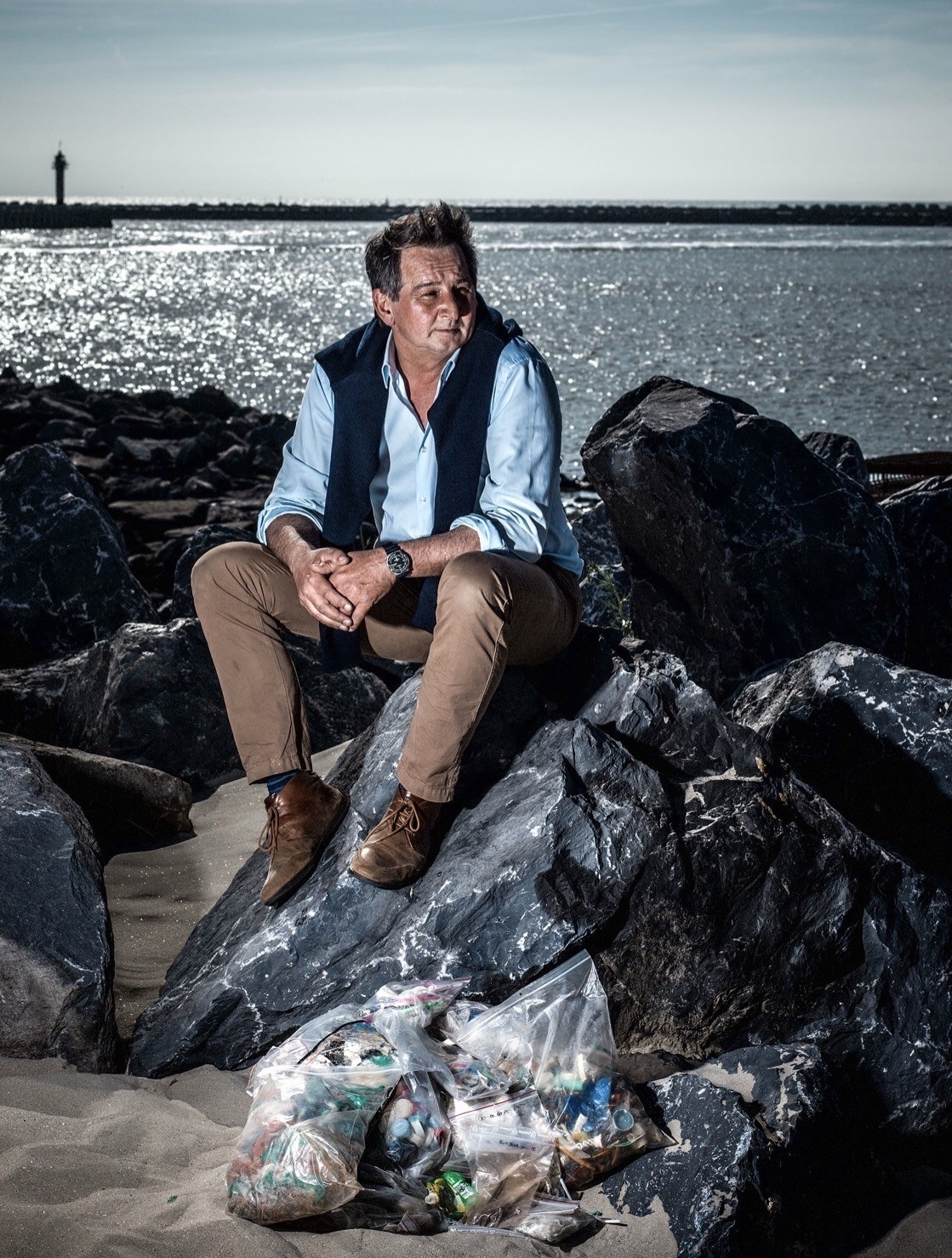 Een formeel geklede man, Colin, zit op een golfbreker naast zakken afval aan de Noordzee.