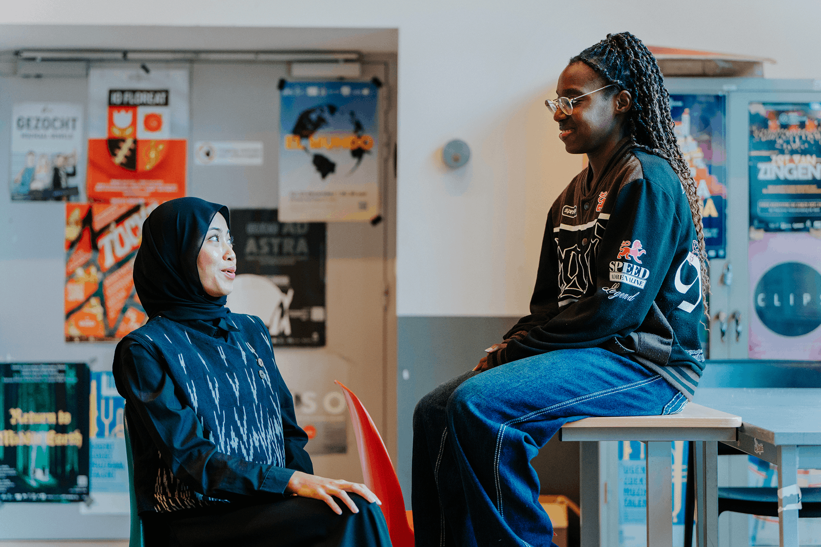 Aini en Ariana poseren en glimlachen naar elkaar op een tafel en stoelen in de hal van een campus.