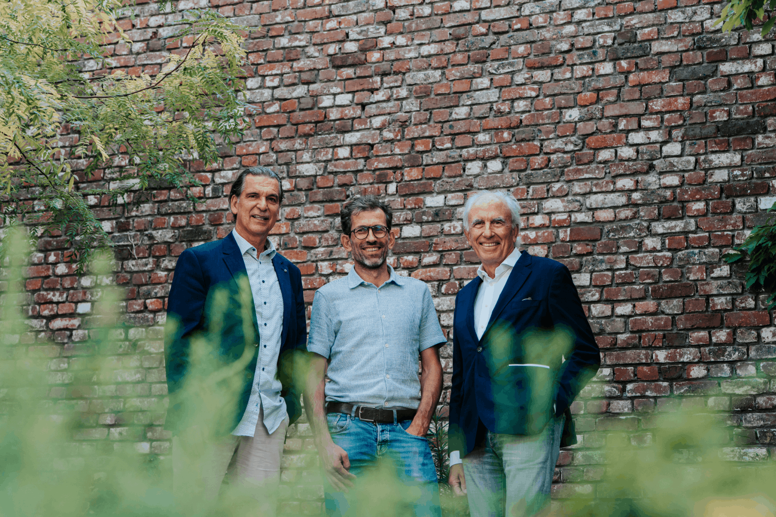 Drie formeel geklede mannen poseren voor een bakstenen muur in een tuin.