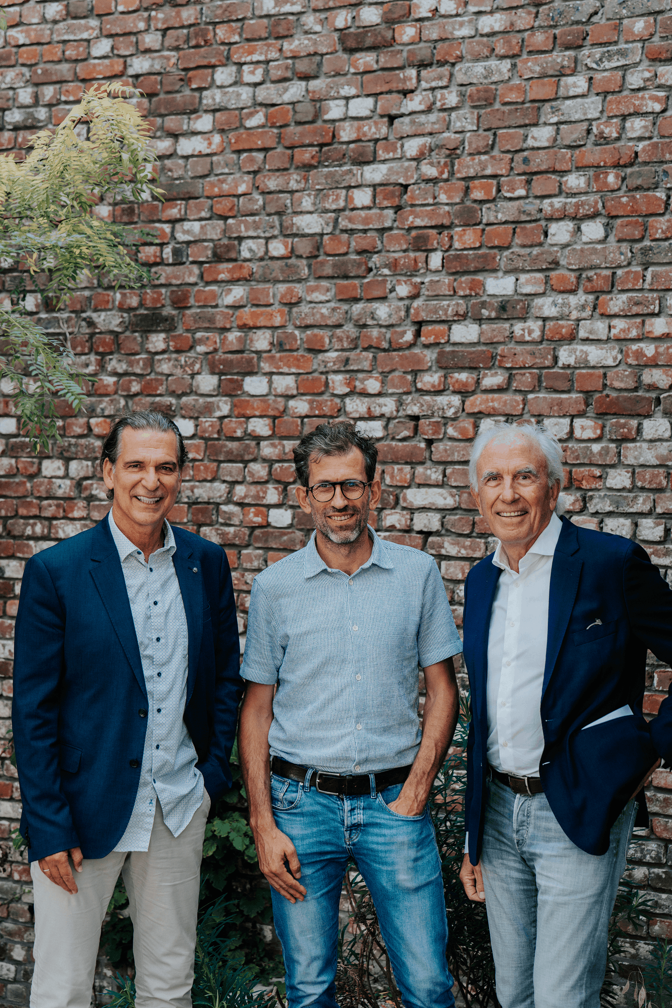 Drie formeel geklede mannen poseren voor een bakstenen muur.