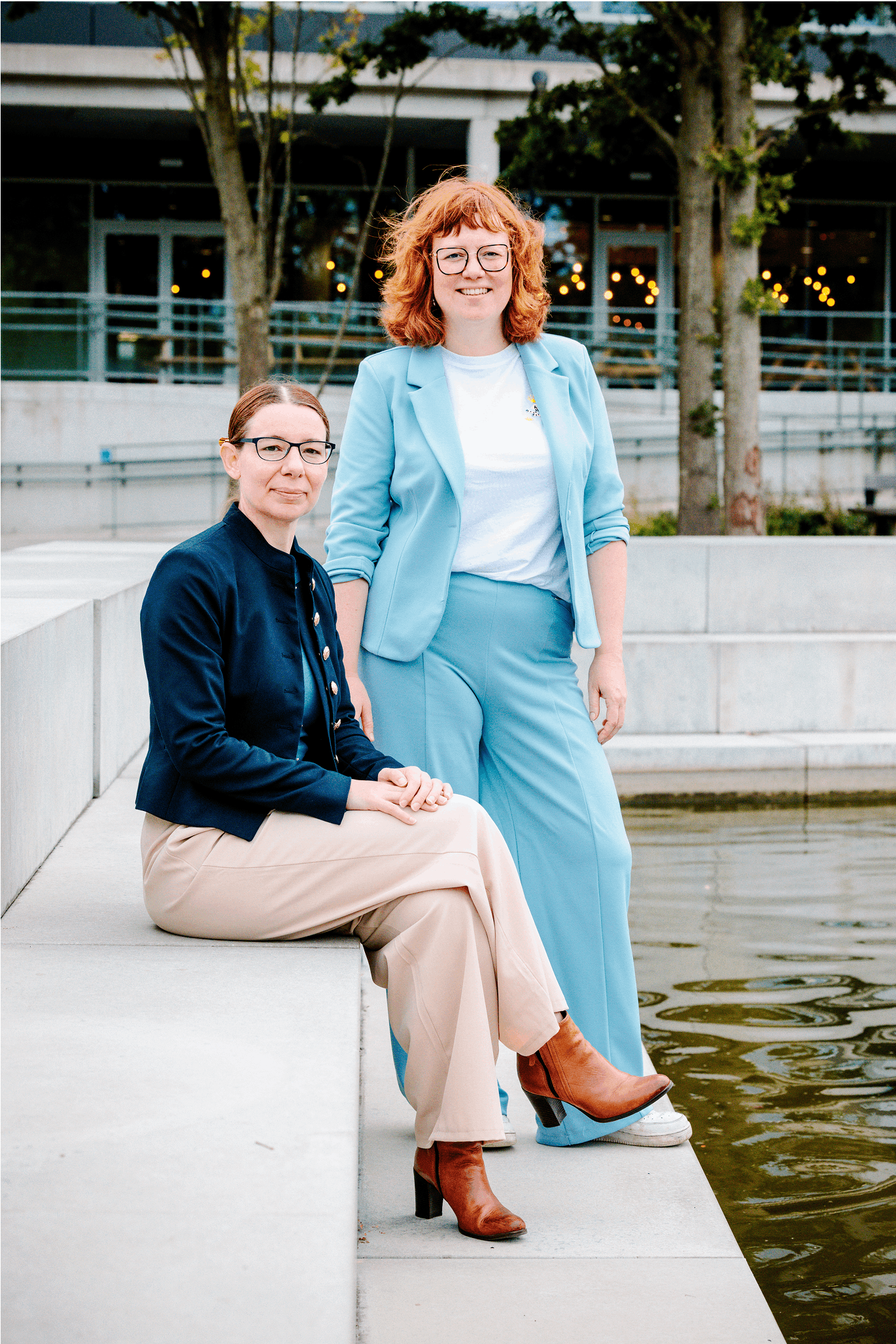 Elisabeth en Wendy zitten op een betonnen bank aan de vijver van een modern gebouw.