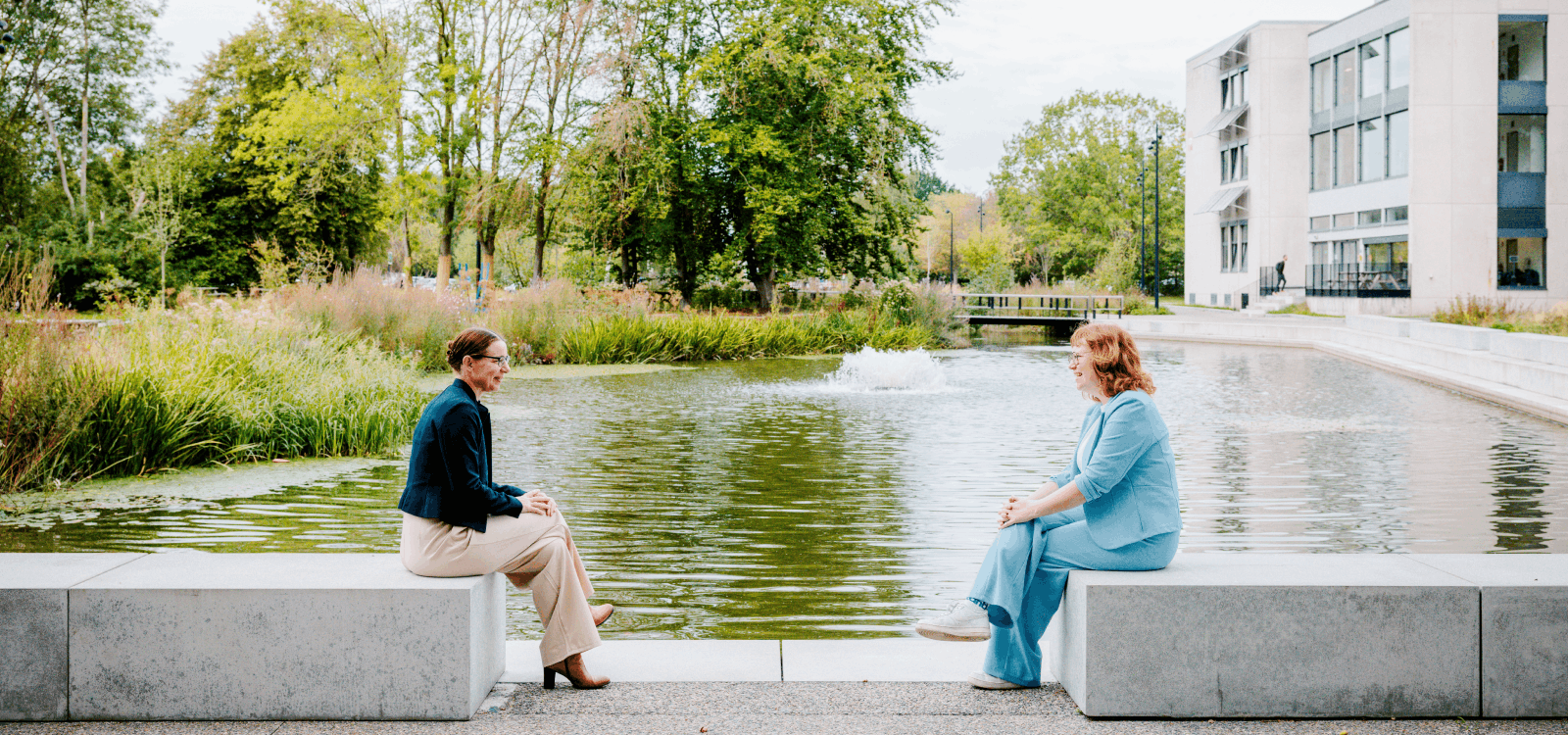 Elisabeth en Wendy zitten op tegenover elkaar op een betonnen bank aan een vijver.