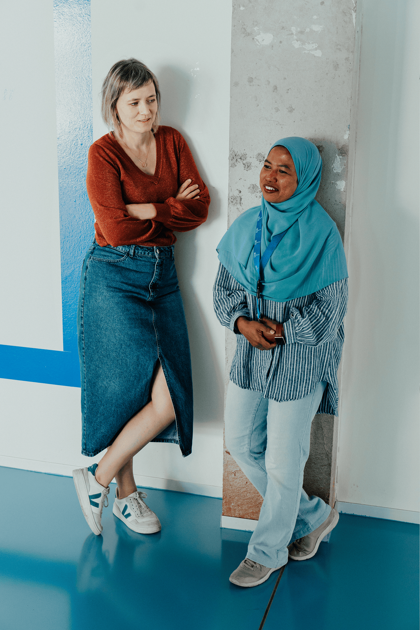 Ellen en Tina leunen tegen de muur in een hal en lachen.