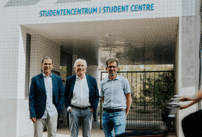 Drie formeel geklede mannen poseren voor het studentencentrum van UGent.