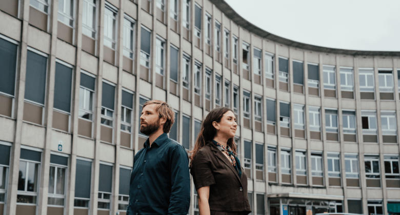 Thomas en Dayana staan rug aan rug voor de buitengevel van een campus.