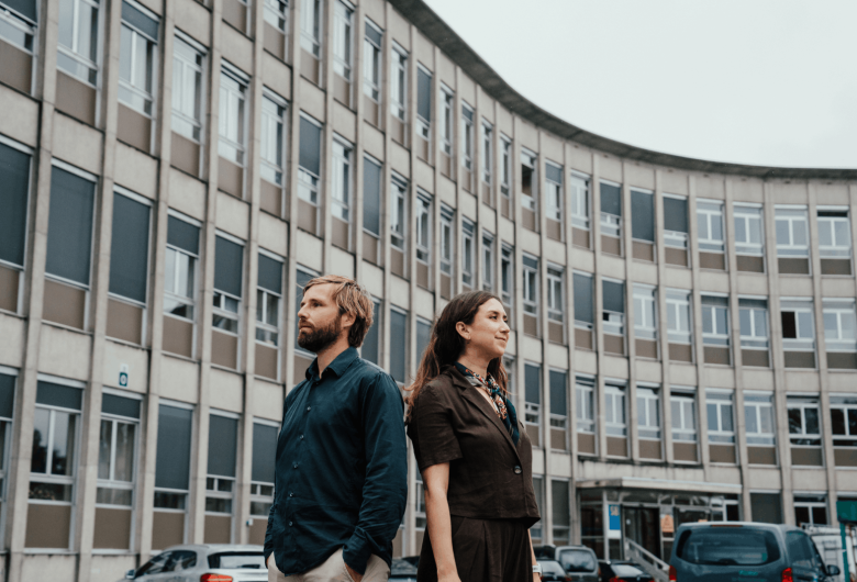 Thomas en Dayana staan rug aan rug voor de buitengevel van een campus.